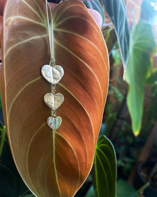 A silver pendant of 3 chain of hearts leaves hanging from a fine silver chain in front of an amber coloured velvet leaf and greenery in the background