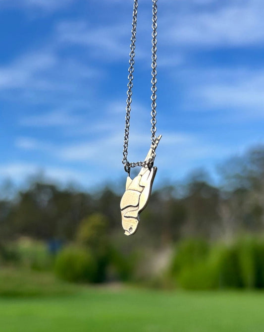 a silver pendant of a Rainbow lorikeet bird hanging upside down, its legs hanging from a fine silver chain in front of a blurred background of green grass and blue sky.