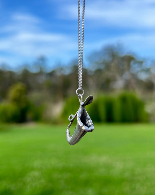 a silver pendant of a nepenthes 'pitcher plant' on a fine silver chain in front of a blurred background of green grass and blue sky.